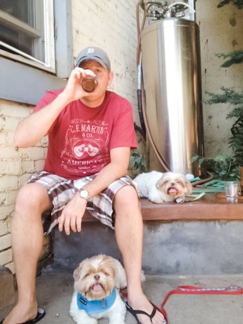 Man sitting on a low stone wall drinking a beer with a small dog next to him and another larger dog at his feet