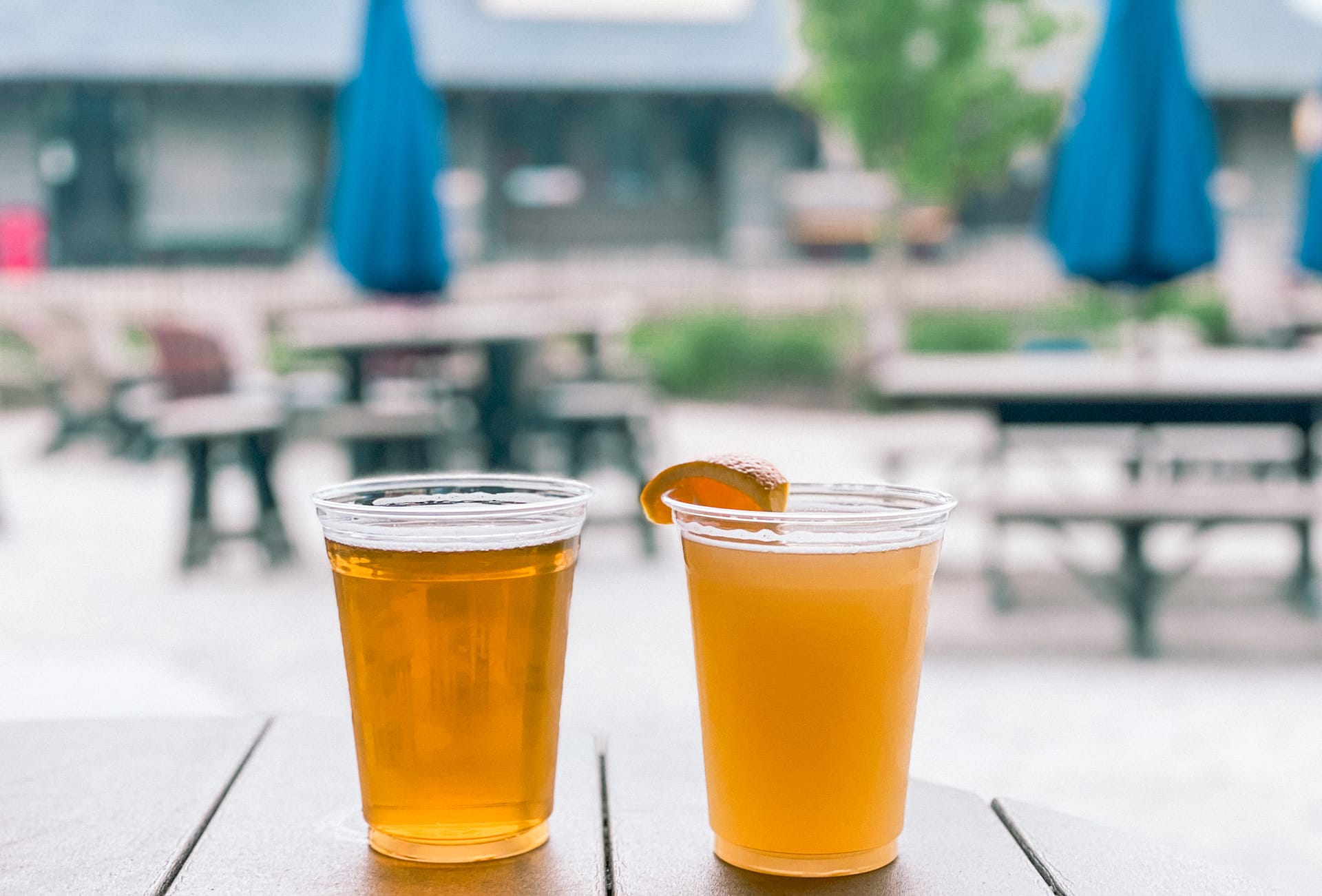 Two cups of beer, one with an orange slice at the top, beer garden in background